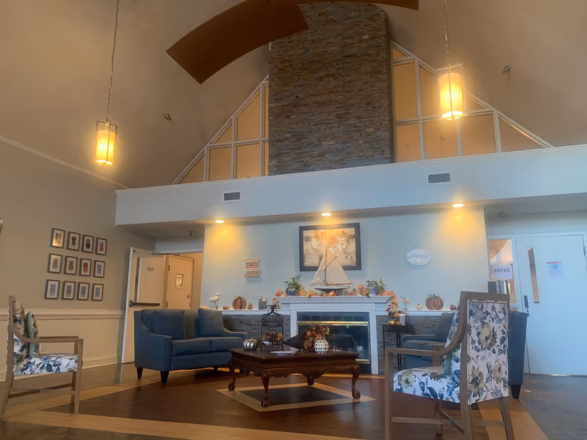 A cozy living room area with a high vaulted ceiling and a large stone fireplace. The room features a blue loveseat, floral upholstered chairs, a wooden coffee table with decorative items, and warm pendant lights hanging from the ceiling. There are framed pictures on the wall to the left and doors labeled 'EXIT' and 'ENTER' on the right side.