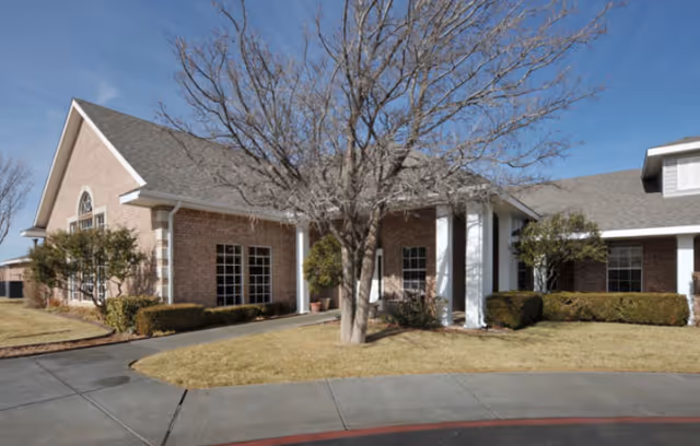 Exterior view of a single-story brick building with white columns at the entrance, leafless trees, trimmed bushes, and a clear blue sky.