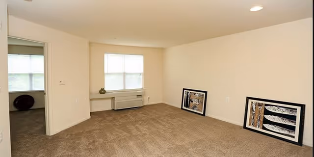 Empty living room with beige carpet and cream-colored walls. Two framed artworks are resting against the wall on the floor. A window with blinds is centered on the far wall above a heating/cooling unit. An open doorway leads to another room with a window and carpeted floor.