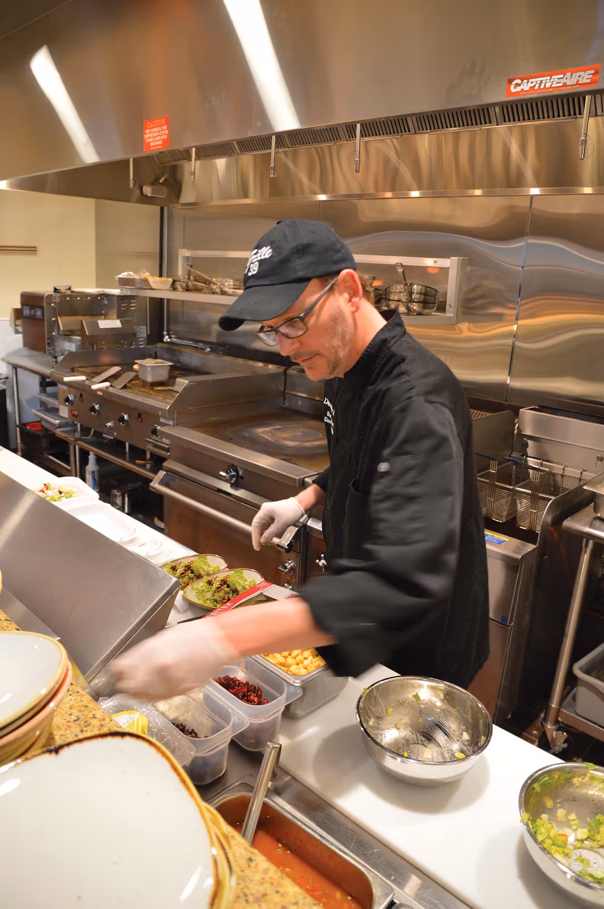 A chef wearing a black uniform, glasses, and a black cap is preparing food in a commercial kitchen. He is adding ingredients to plates on a counter with various containers of food and bowls around him. The kitchen has stainless steel appliances and a large ventilation hood above the cooking area.
