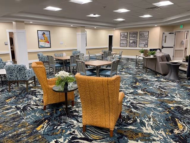 A spacious common area with patterned carpet, several tables with chairs, and two wicker armchairs around a small round table with a flower arrangement. The walls have framed pictures and there is a sofa with a side table and plants near the back. The ceiling has recessed lighting.