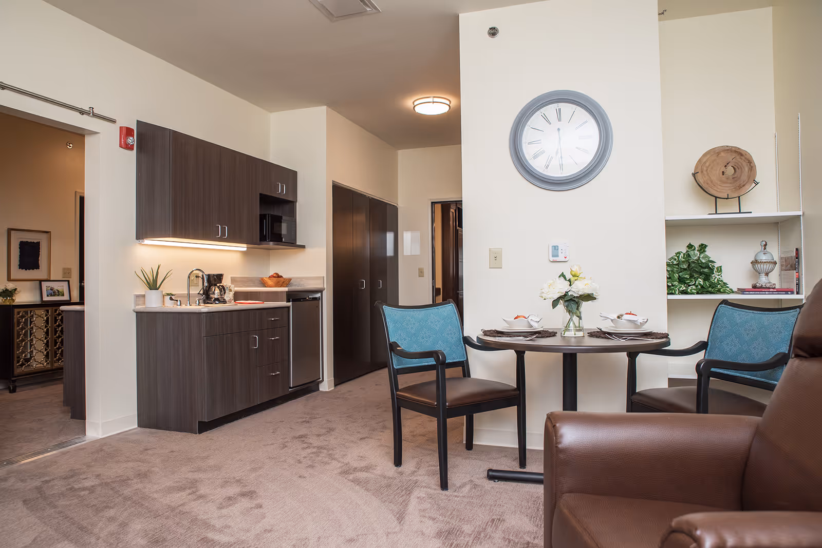 Interior view of a senior living facility apartment featuring a small kitchenette with dark wood cabinets, a sink, coffee maker, and microwave. Adjacent to the kitchenette is a round dining table set for two with blue cushioned chairs. A large wall clock is mounted on a white wall above the table. The room has beige carpet and neutral-colored walls, with a brown leather armchair partially visible in the foreground. Shelves with decorative items and plants are on the right side.