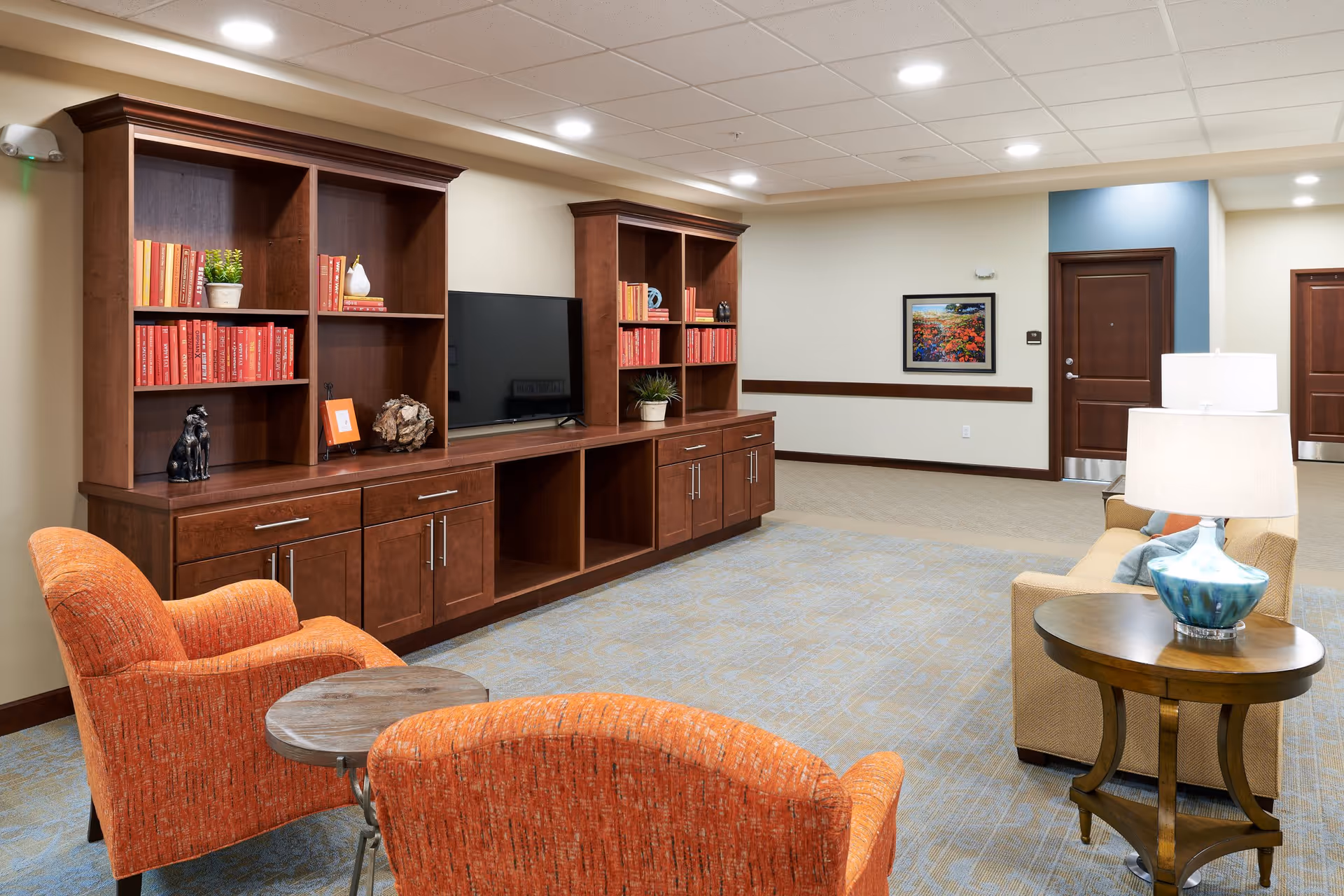 A comfortable senior living common room with orange armchairs, a sofa, wooden built-in shelving and a TV.