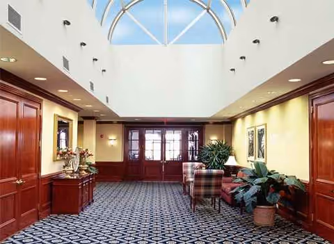 Spacious lobby with wood paneling, patterned carpet, plants, and seating beneath a skylit ceiling.