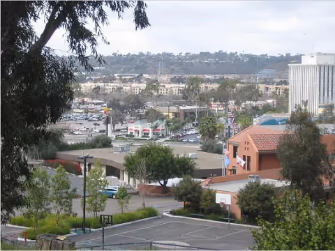View overlooking a cityscape with various buildings, trees, and a basketball court in the foreground. The scene includes a mix of commercial and residential structures under a clear sky.