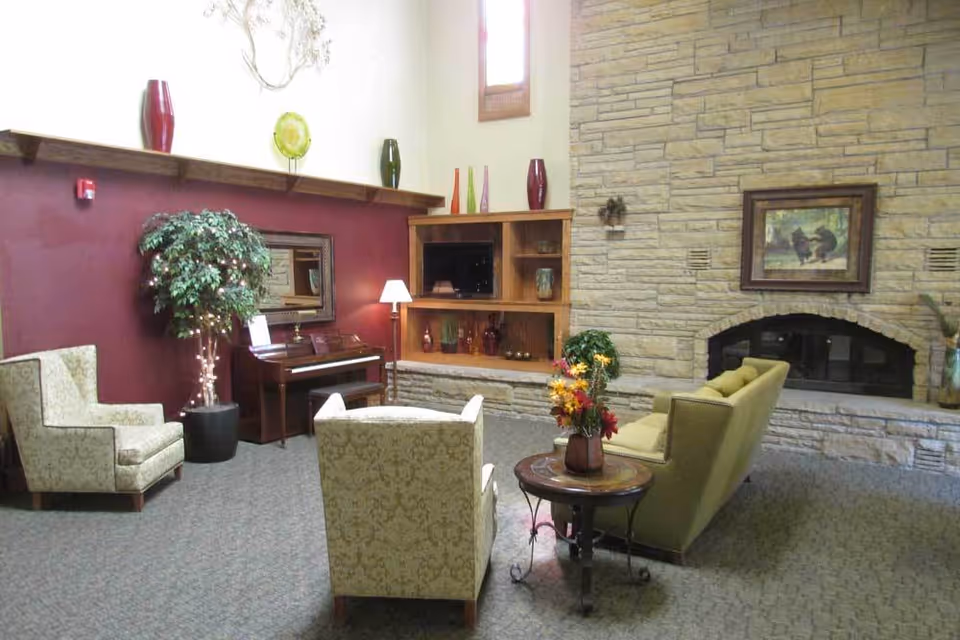 Seating area with upholstered chairs, a sofa, piano, built-in shelving, and a stone fireplace.