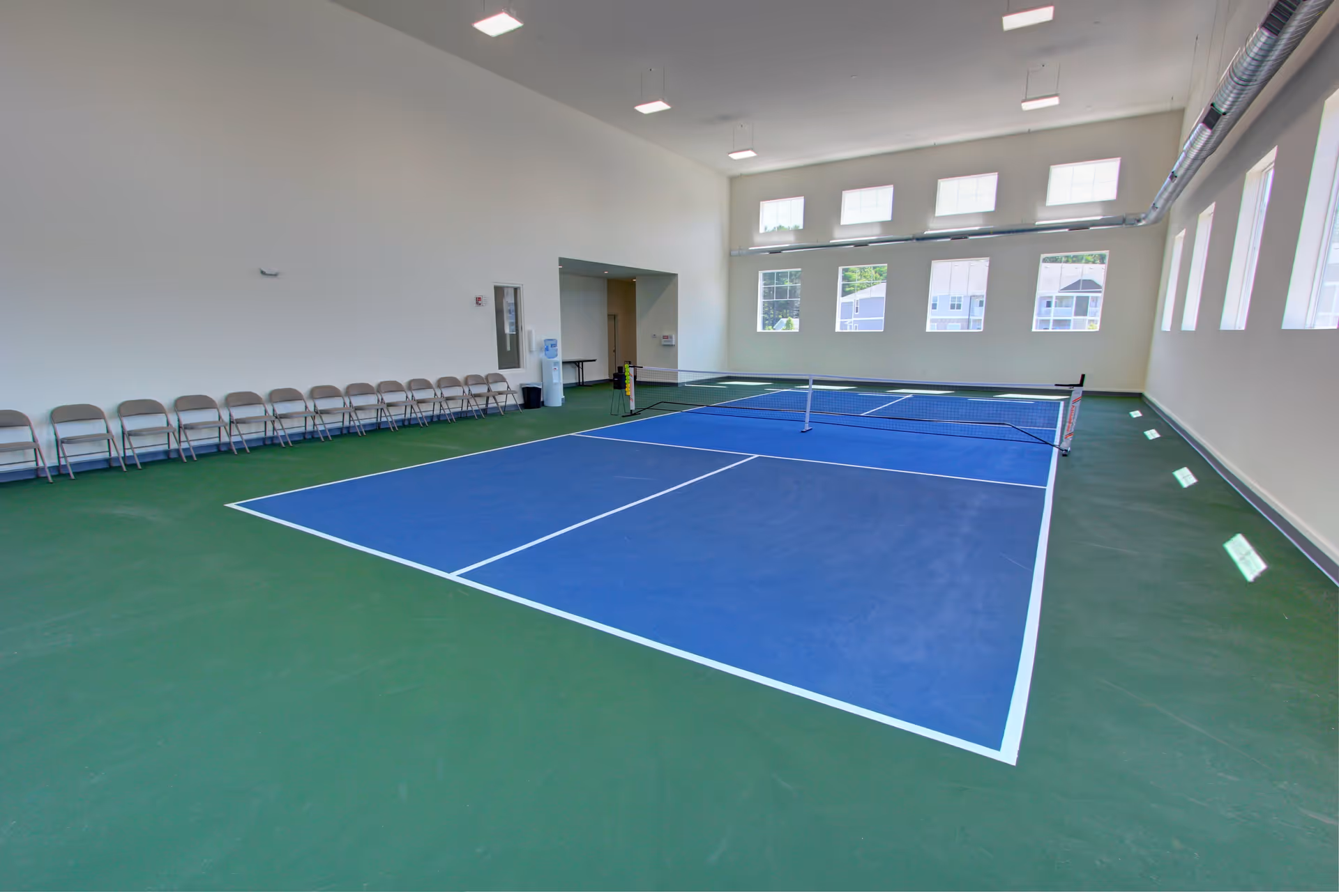 Indoor pickleball court with blue playing area and green surrounding floor, lined with folding chairs along one wall, large windows letting in natural light, and exposed ductwork on the ceiling.