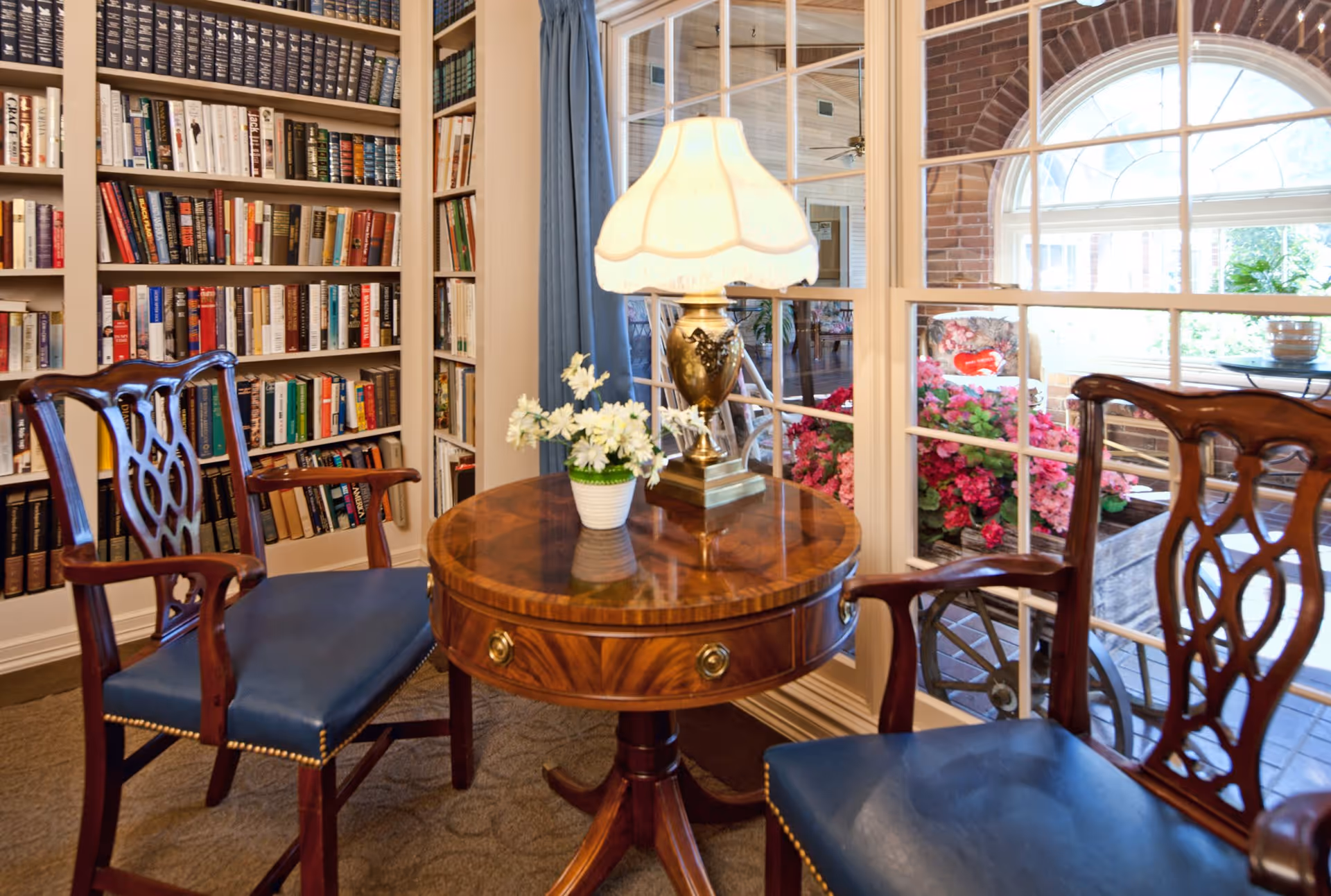 A cozy interior reading nook with two wooden chairs featuring blue cushions around a round wooden table. On the table is a decorative lamp with a white shade and a small white pot with white flowers. Behind the table is a large window with multiple panes, showing a view of a bright outdoor area with pink flowers and brick walls. To the left, there is a tall bookshelf filled with books.