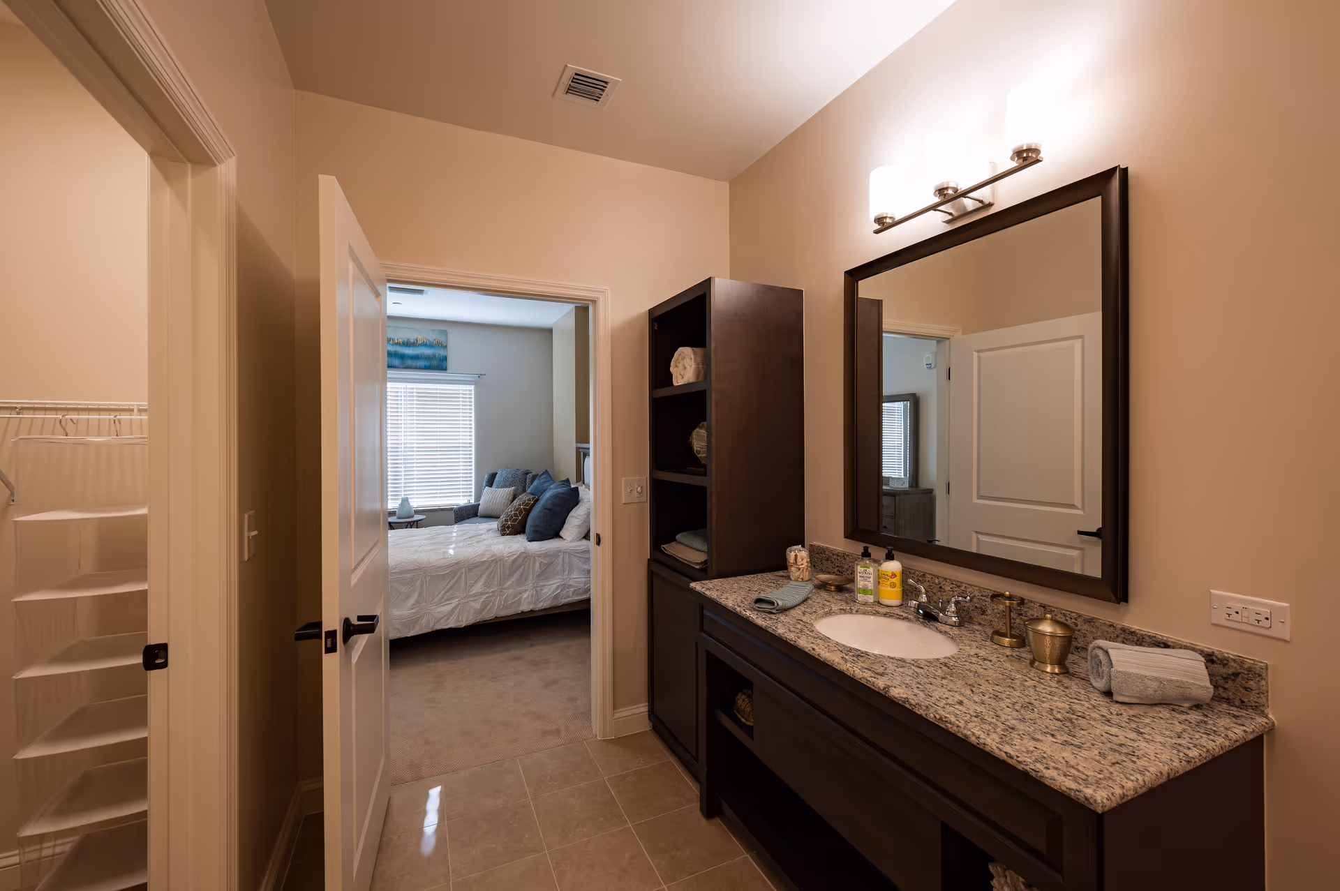 View of a bathroom with a granite countertop, sink, and a large mirror above. There is a dark wood cabinet with shelves holding towels next to the sink. The bathroom door is open, showing a bedroom with a bed, pillows, and a window with blinds.