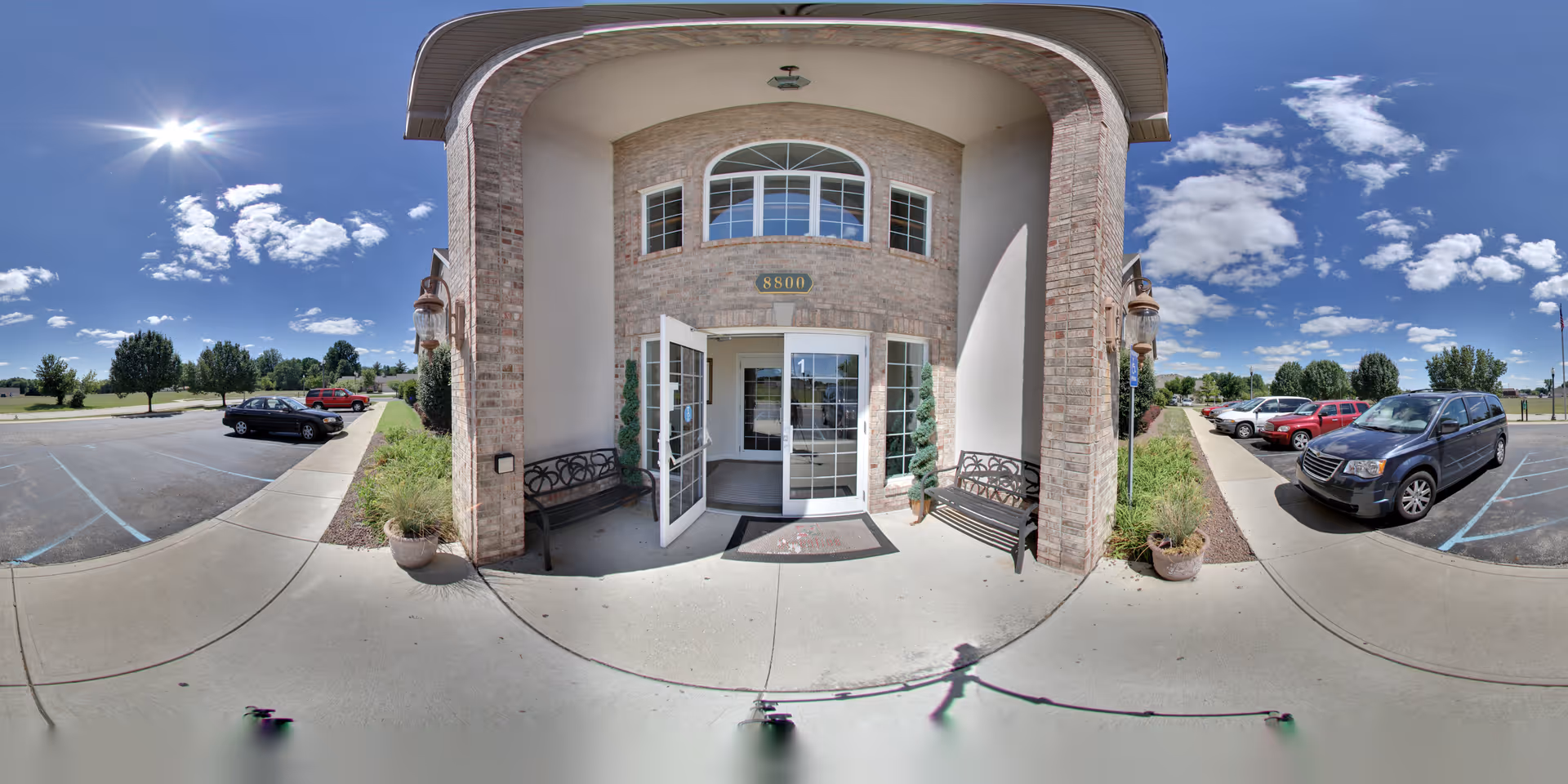 Entrance of a building with double glass doors open, flanked by two benches and potted plants. The building has a brick facade with the number 8800 above the door. There is a parking lot on both sides with several parked cars under a bright blue sky with scattered clouds.
