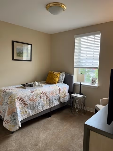 A small bedroom with a single bed covered in a colorful quilt with leaf patterns. There is a yellow pillow and a gray pillow on the bed. Next to the bed is a round metal side table with a lamp and a small framed photo. A window with white blinds is behind the table. A framed picture hangs on the beige wall above the bed. The room has beige carpet and neutral-colored walls.