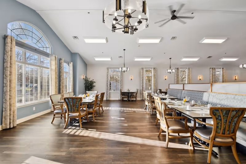 Bright and spacious dining room with large windows allowing natural light to fill the space. The room features wooden floors, multiple dining tables with chairs, cushioned bench seating along one wall, and modern chandeliers hanging from the ceiling. Light blue walls and patterned curtains add a cozy atmosphere.