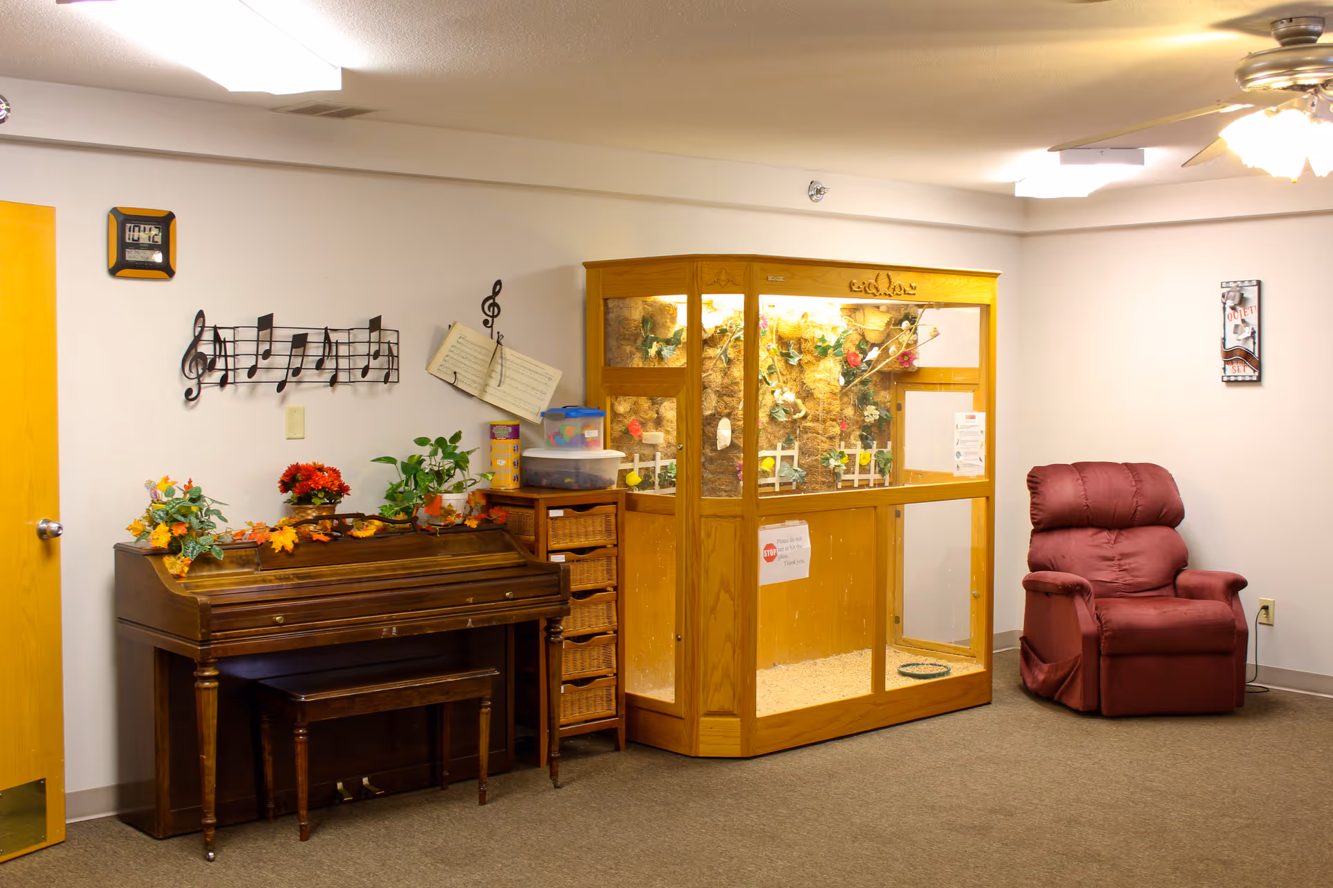 A cozy room with a wooden piano decorated with artificial flowers and plants, a wooden cabinet with wicker baskets, a large wooden enclosure with plants and decorations inside, and a red recliner chair. The walls are light-colored with musical note decorations and a clock.