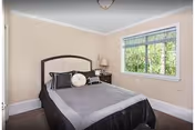 Small bedroom with a bed, headboard, nightstand with a lamp, and a window showing greenery outside.