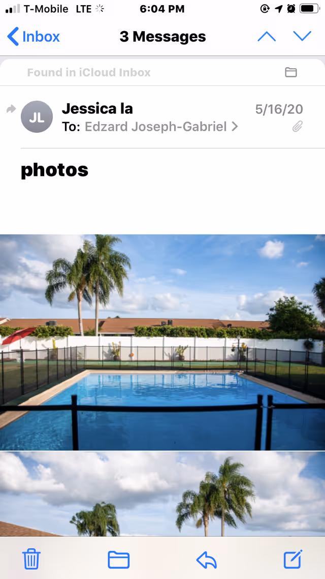 A fenced outdoor swimming pool with clear blue water, surrounded by a grassy area and palm trees under a partly cloudy sky.