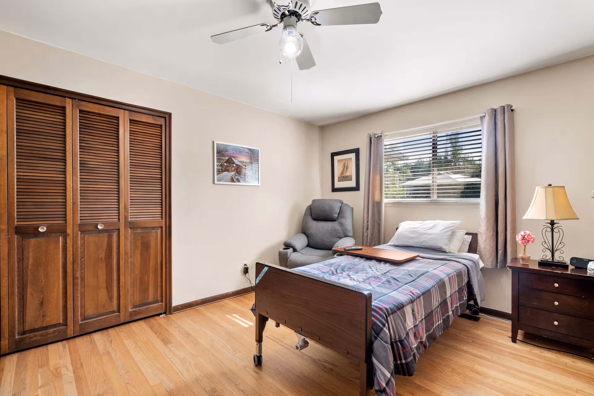 A simple bedroom with a single bed covered in a plaid blanket, a gray recliner chair next to the bed, a wooden nightstand with a lamp and a small flower vase, a window with blinds and curtains, a ceiling fan with a light, and a wooden closet with louvered doors.
