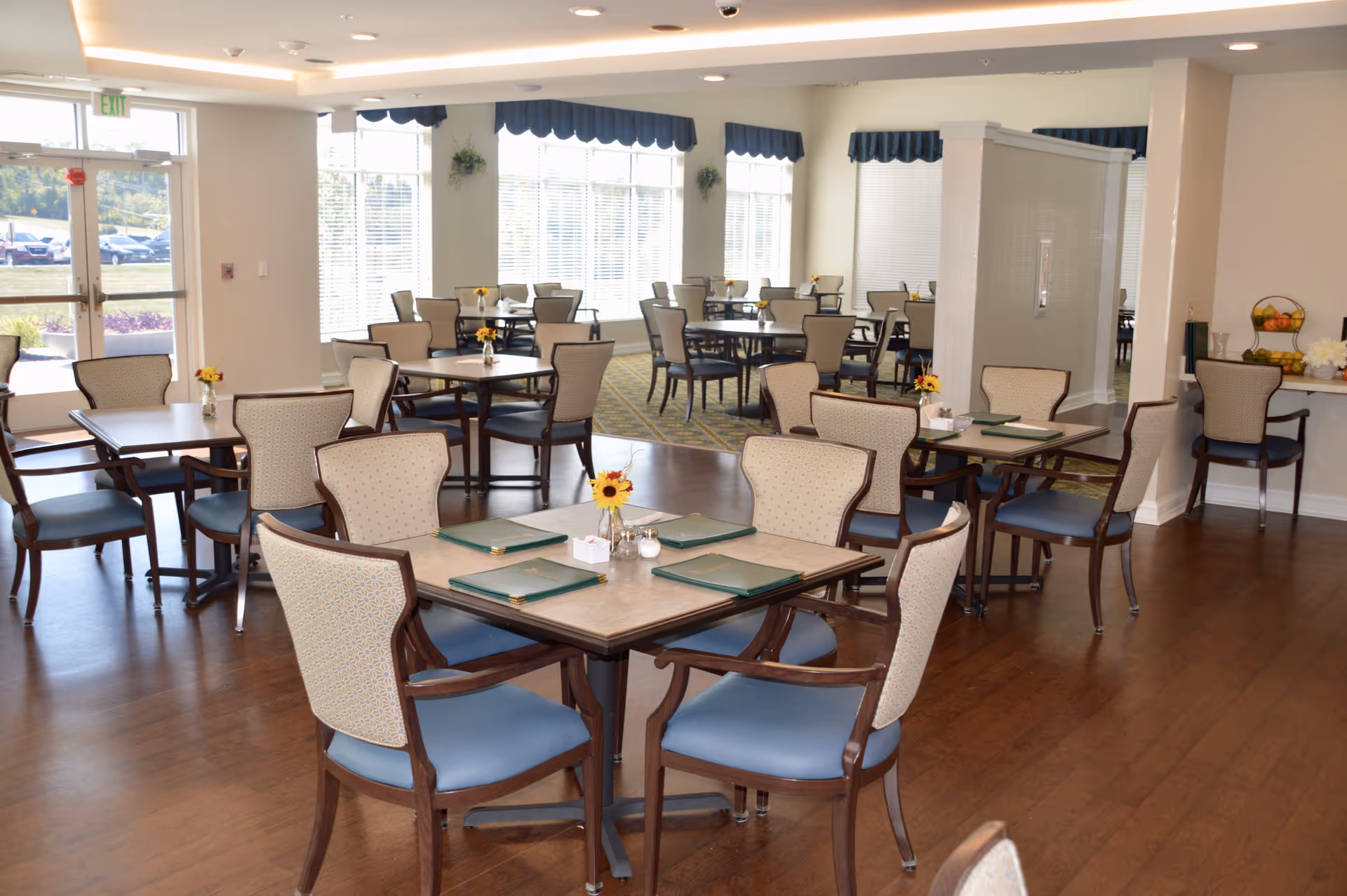 Well-lit dining room with multiple tables and chairs set with place mats and small floral centerpieces.