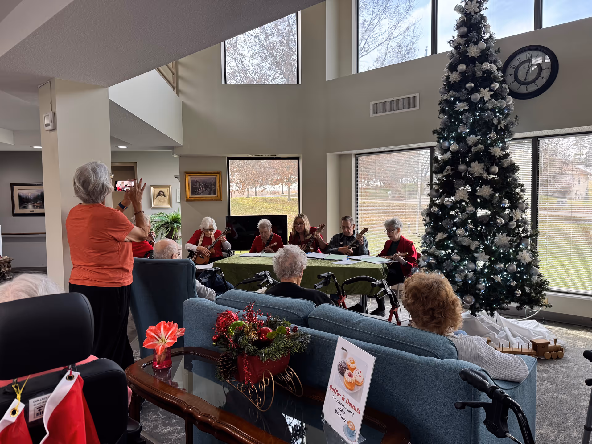 A group of elderly people seated in a living room area with large windows and a tall decorated Christmas tree. Some are playing string instruments while others watch. One woman is standing and taking a photo with a smartphone. The room is decorated with holiday-themed items including a small floral centerpiece on a glass table.