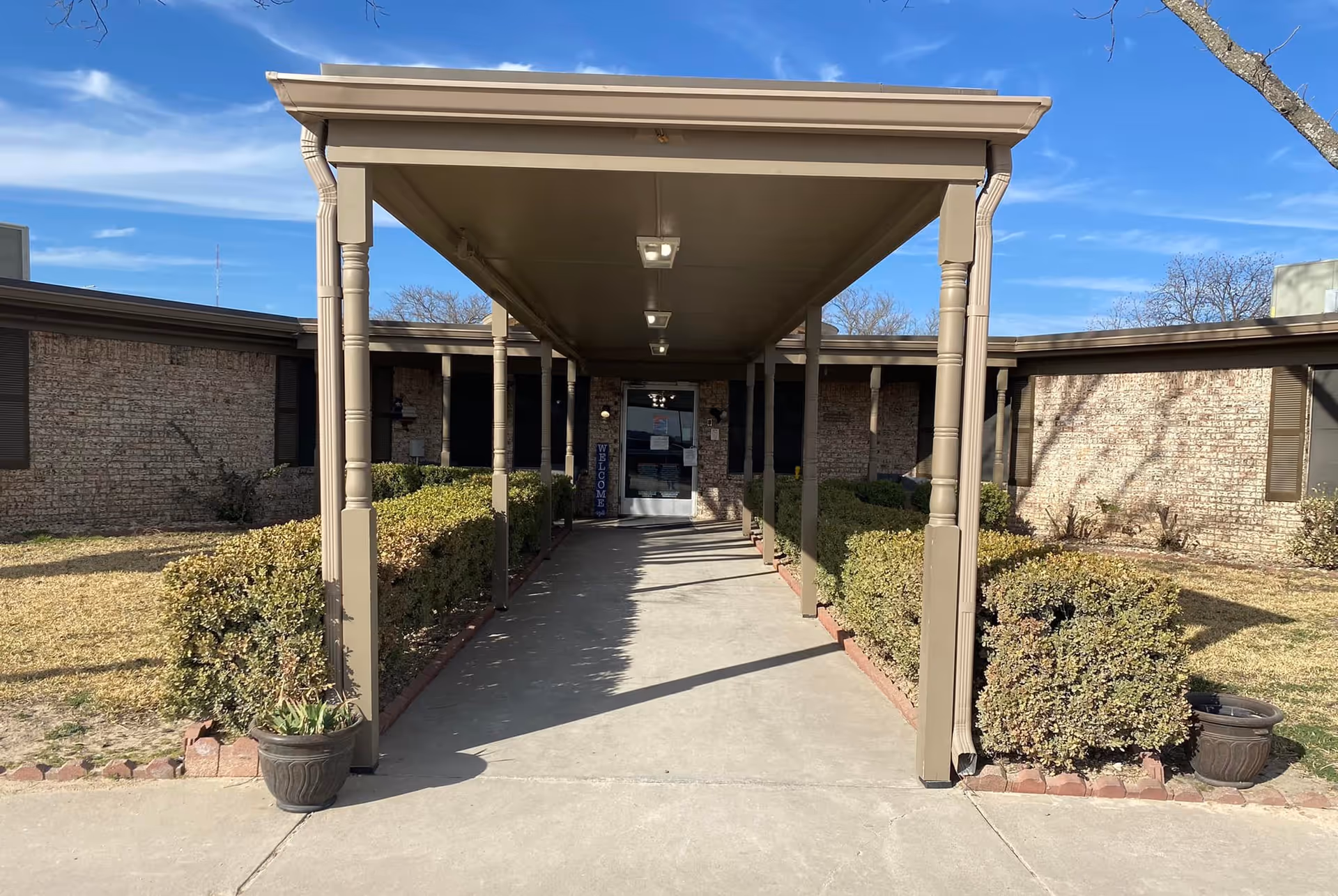 Covered walkway leading to the entrance of a single-story brick building with trimmed bushes on both sides under a clear blue sky.