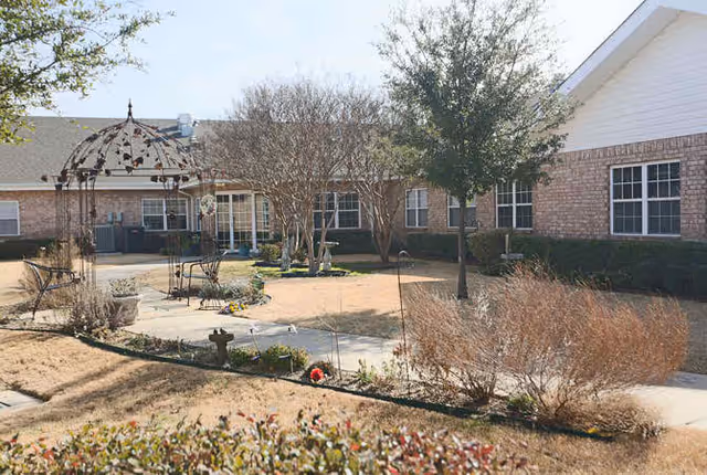 Outdoor courtyard area at Mercer Terrace featuring a paved walkway, a decorative metal gazebo with chairs underneath, leafless trees, bushes, and a brick building with multiple windows in the background.