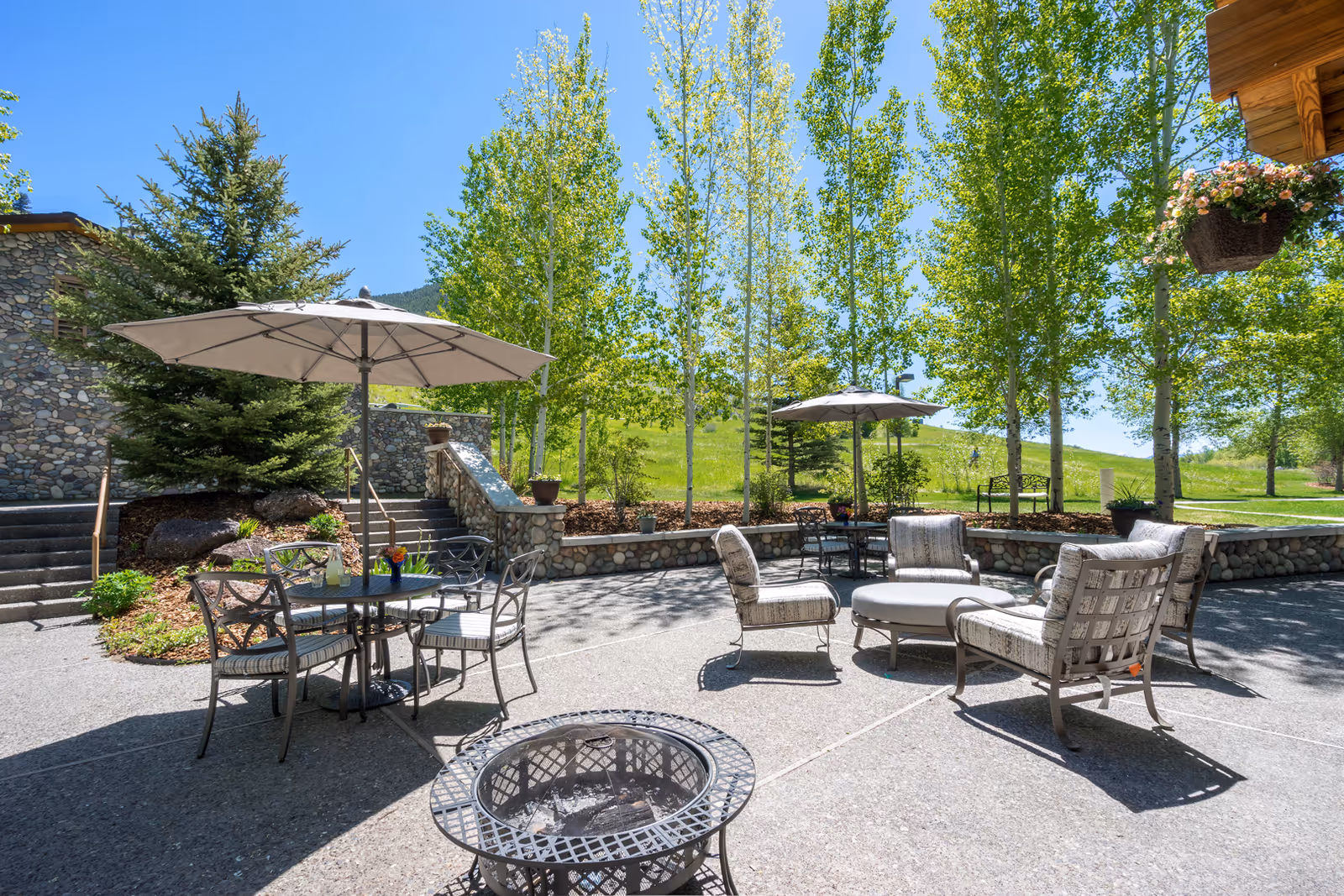 Outdoor patio area with metal chairs and tables under large umbrellas, cushioned lounge chairs arranged around a fire pit, surrounded by green trees and a stone wall, under a clear blue sky.