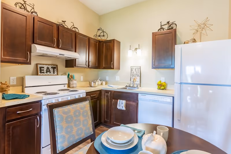A bright kitchen with dark wood cabinets, a white stove, dishwasher, and refrigerator. The countertop has a bowl of fruit, kitchen utensils, and decorative signs that say 'EAT' and 'EAT DRINK & GIVE THANKS'. A round dining table in the foreground is set with white dishes, cups, and a teal placemat.