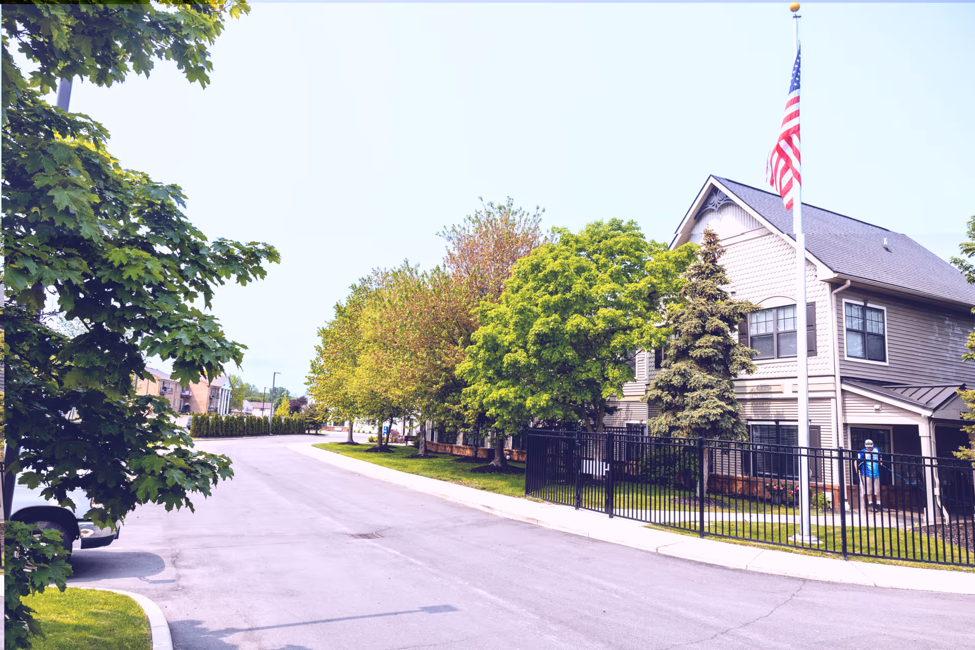Exterior view of a senior living facility with a paved road, green trees, and a two-story building with beige siding and an American flag on a flagpole in front. A person wearing a blue shirt and hat is visible near the building entrance.