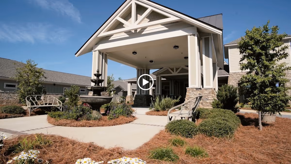 Front entrance of an assisted living building with a covered porte-cochere, central fountain, benches, and landscaped walkways.