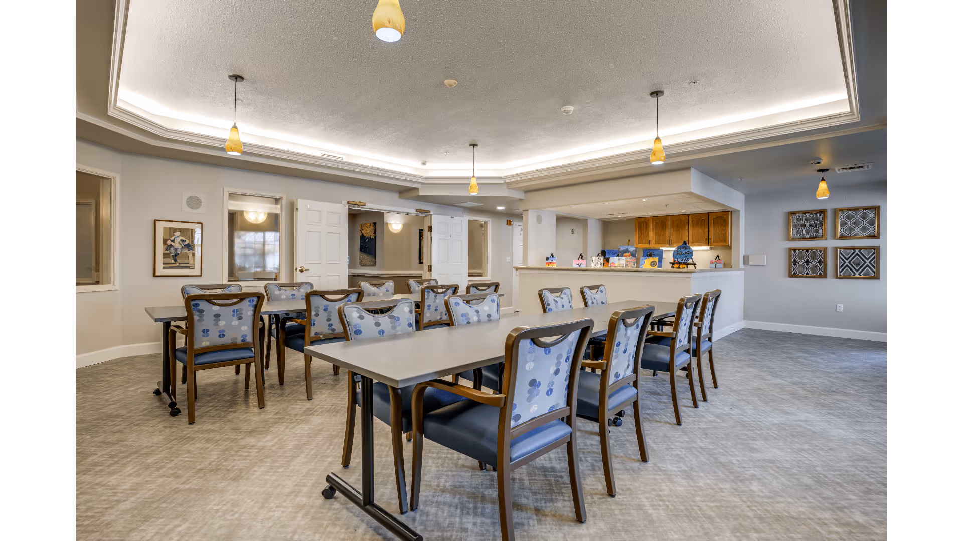 A spacious dining room with two long tables surrounded by chairs with blue patterned upholstery. The ceiling features recessed lighting and hanging pendant lights with yellow shades. The room has light-colored walls and flooring, with framed artwork on the walls and a counter area with wooden cabinets in the background.