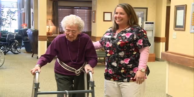 An elderly woman using a walker is being assisted by a smiling female caregiver in a hallway of a senior living facility.