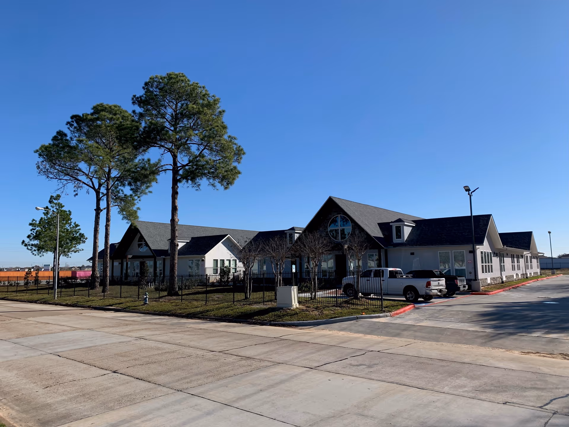 Exterior view of Silvercrest Assisted Living and Memory Care building on a clear day, showing a single-story structure with a dark roof, large windows, and a fenced grassy area with tall pine trees. Several parked vehicles are visible in the parking lot adjacent to the building.