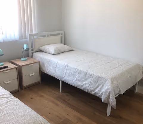 A small bedroom with two beds, a nightstand, lamp, and window blinds.