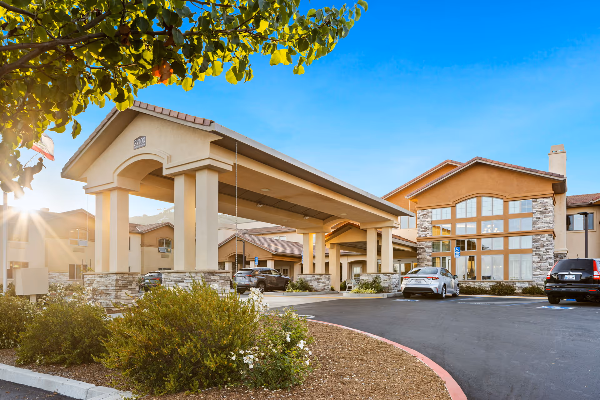 Exterior view of Ivy Park At Murrieta senior living facility with a covered entrance, several parked cars, landscaped bushes, and a clear blue sky with sunlight filtering through tree leaves.