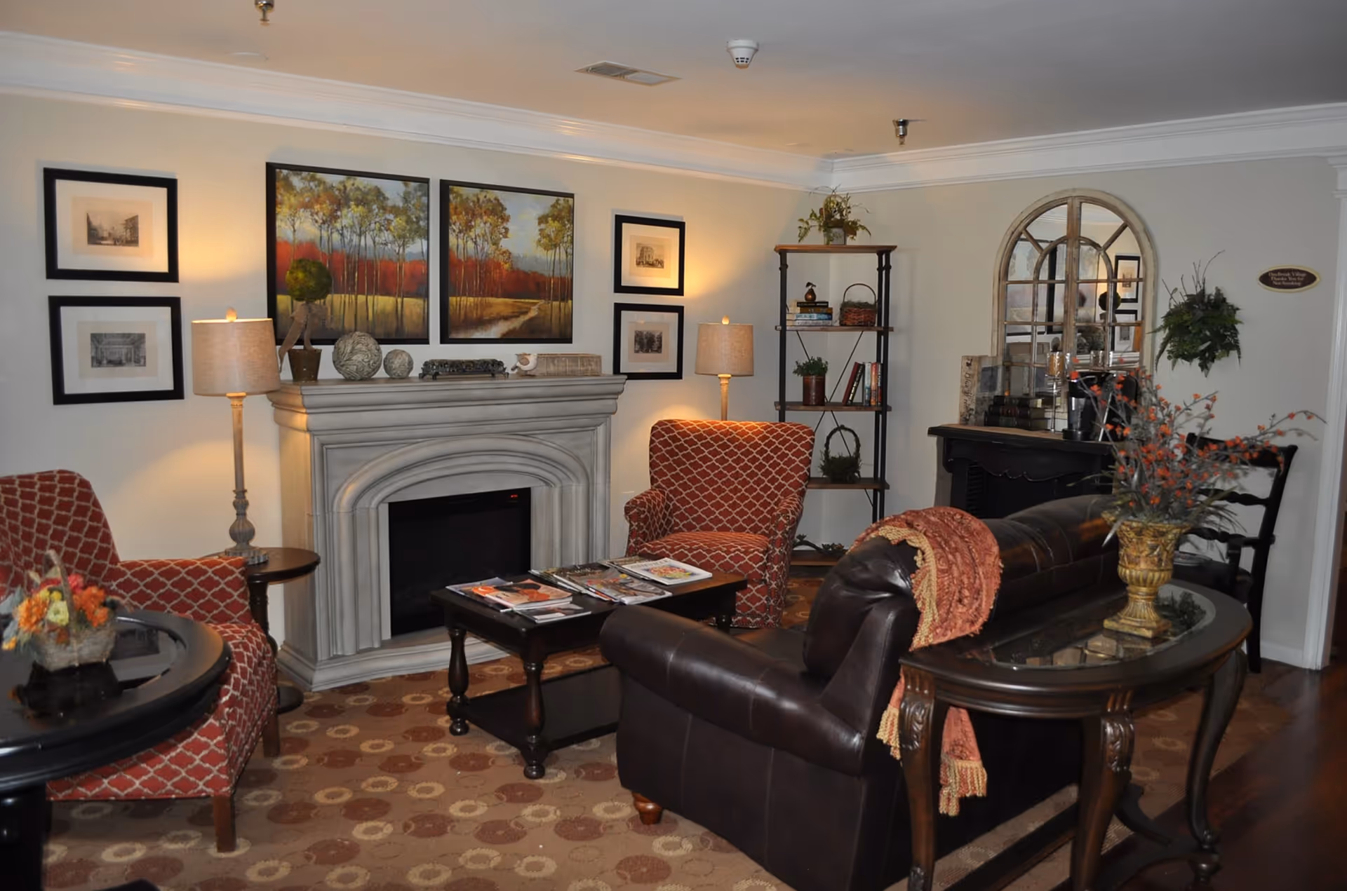 Cozy living room with a dark leather sofa, two red patterned armchairs, a coffee table with magazines, a fireplace with decorative items on the mantel, framed artwork on the walls, two lamps, a bookshelf with plants and books, and a console table with a floral arrangement and a large arched mirror.