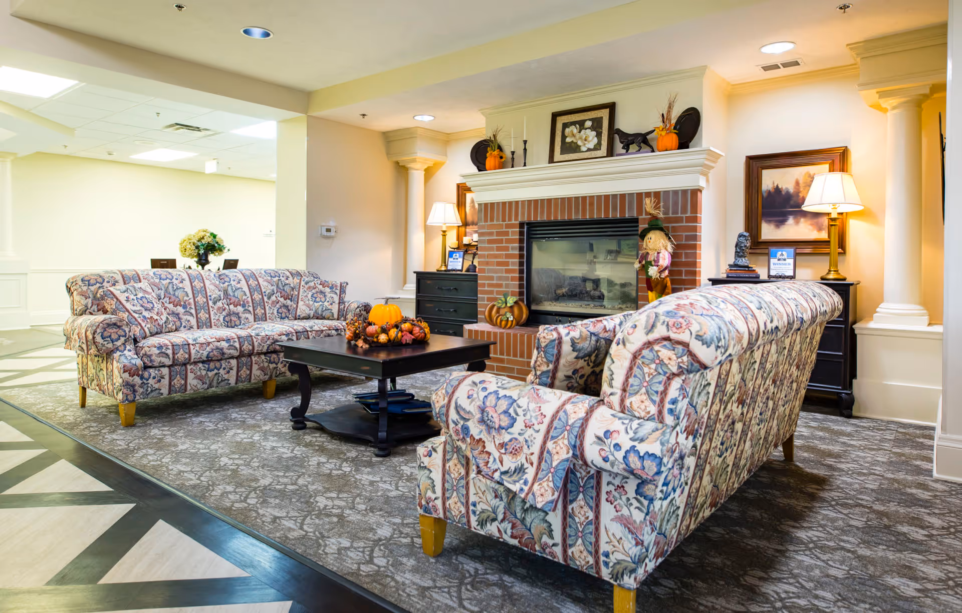 A cozy senior living lounge with two patterned sofas facing a brick fireplace and a coffee table decorated with pumpkins.