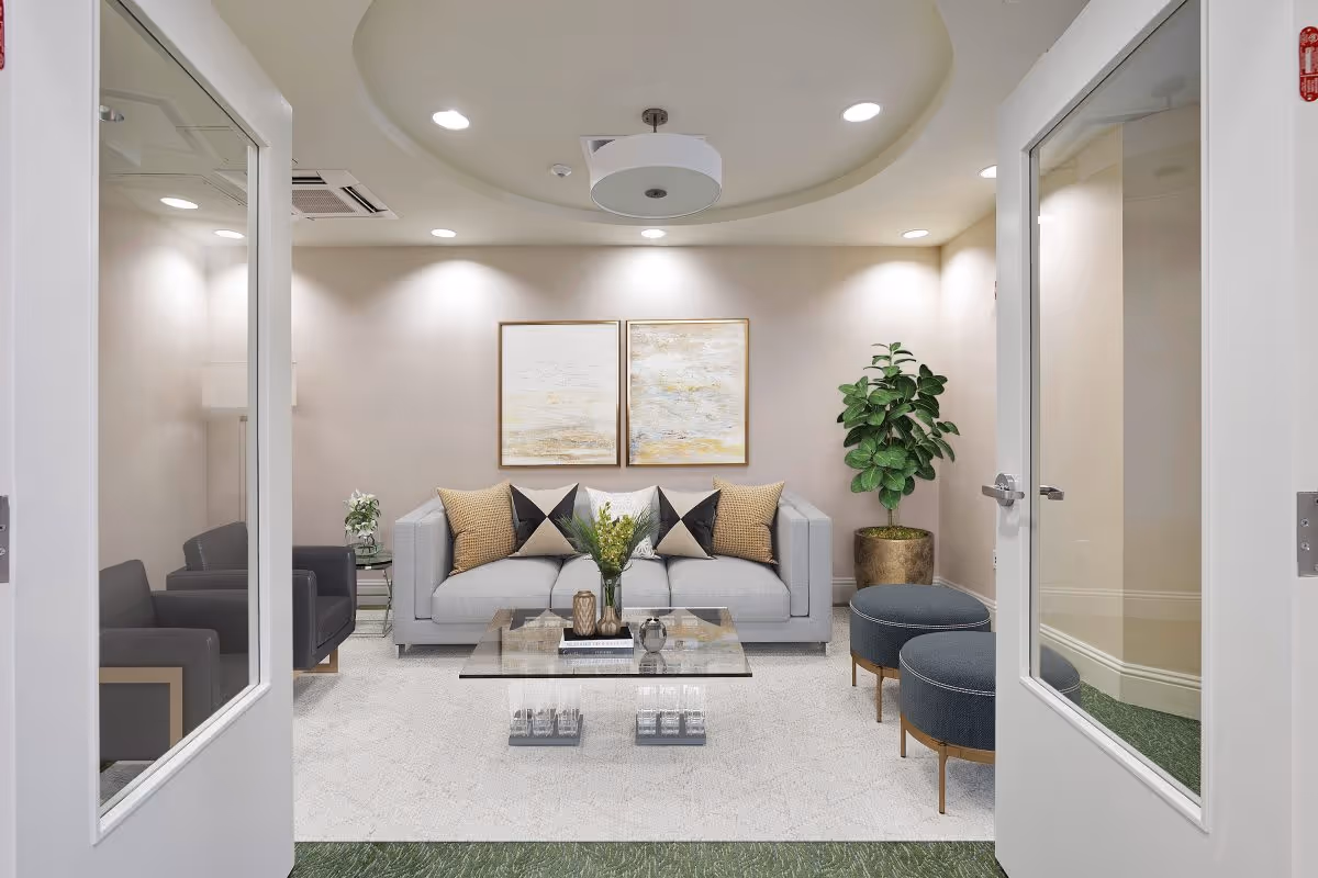 Bright modern lounge viewed through double doors with a sofa, glass coffee table, armchairs, ottomans, artwork, and a potted plant.