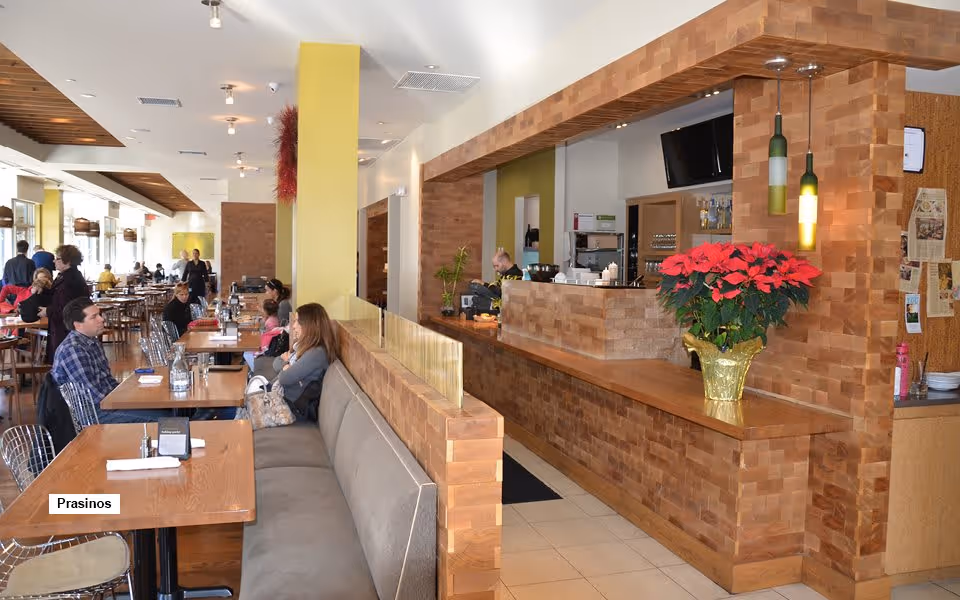 Interior view of a dining area with wooden tables and chairs, a long cushioned bench along one side, and a counter with a poinsettia plant in a gold pot. Several people are seated and engaged in conversation. The space has a warm, inviting atmosphere with wooden accents and pendant lights.