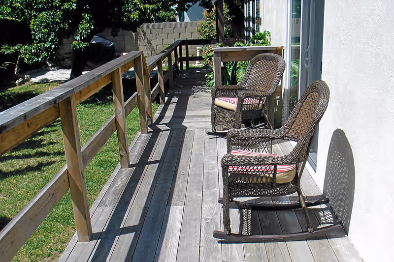 Wooden outdoor deck with two wicker rocking chairs with cushions placed near a white wall and sliding glass door, surrounded by green grass and trees.