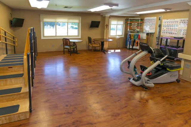 A spacious rehabilitation room with wooden flooring, featuring exercise equipment including a recumbent bike, parallel bars for walking practice, two small tables with chairs near windows, and various therapy tools and charts on the walls.