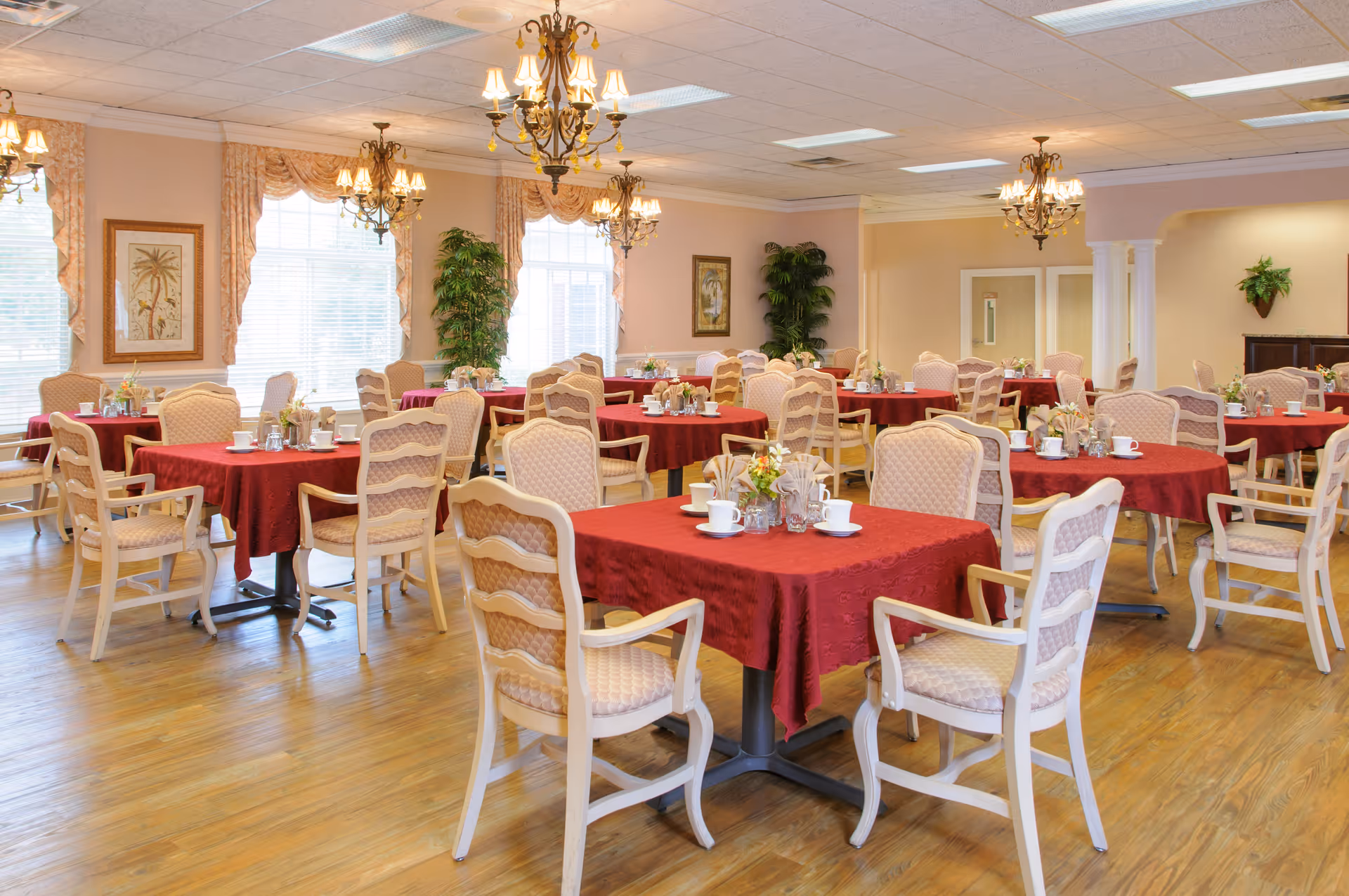 Bright dining room with round tables covered in red tablecloths and beige chairs set for a meal.
