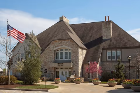 Stone-clad community building entrance with an American flag, landscaped planters, and a curved driveway under a blue sky.