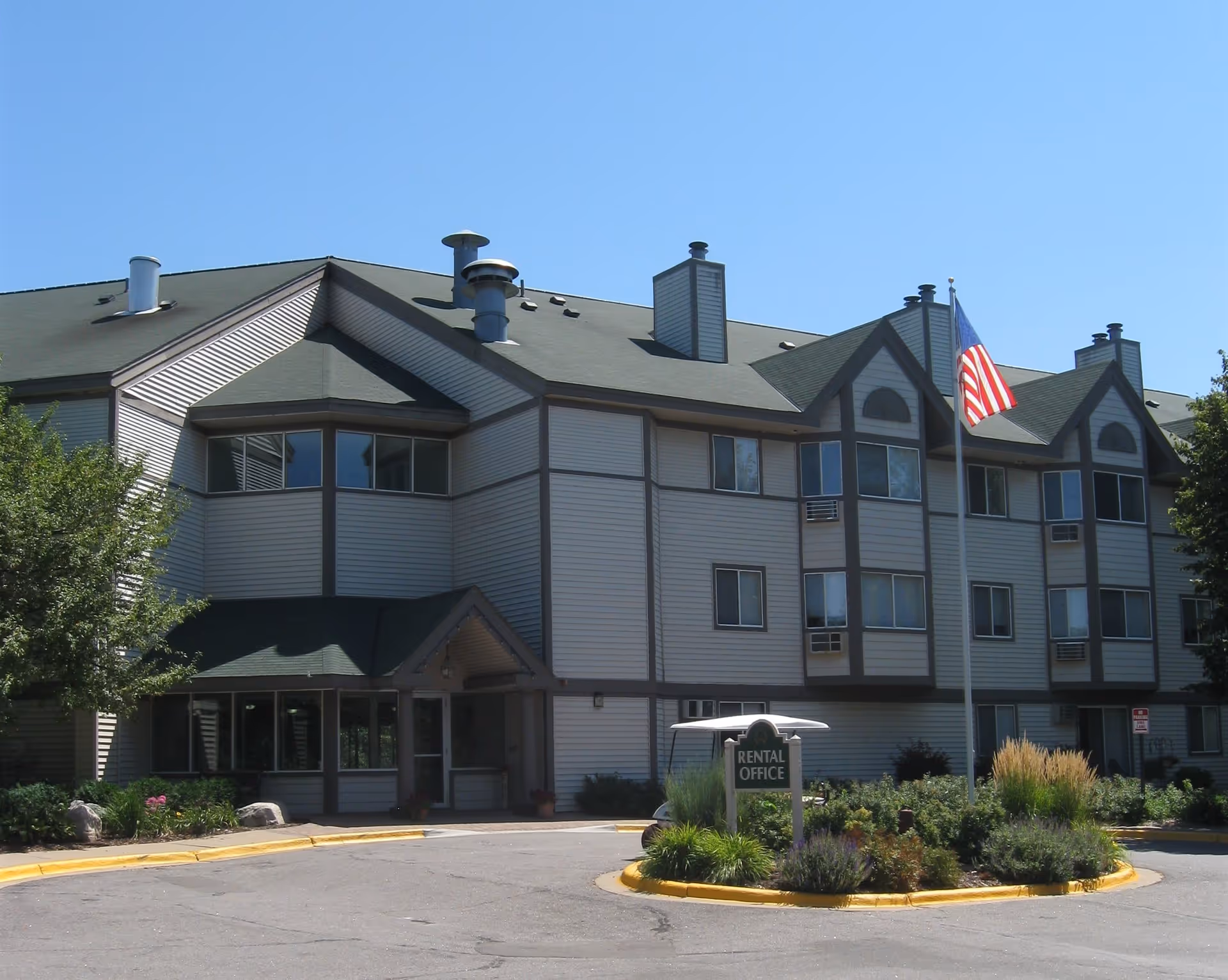 Exterior view of a multi-story senior living facility building with a green roof and beige siding. There is a circular driveway with a landscaped island in the center featuring a sign that reads 'Rental Office' and an American flag on a flagpole. Trees and shrubs surround the building under a clear blue sky.