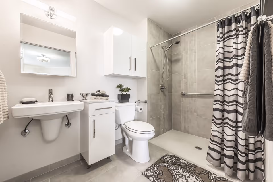 A modern bathroom featuring a wall-mounted sink with a mirror above it, a white cabinet next to a toilet, and a walk-in shower with gray tiled walls and a patterned shower curtain. There are towels hanging on the right side and a decorative rug on the floor.