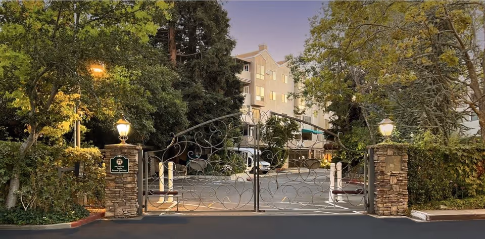 Entrance gate to The Heritage Downtown facility with ornate metal gates, stone pillars topped with lantern-style lights, surrounded by trees and greenery, and a multi-story building visible in the background during dusk.