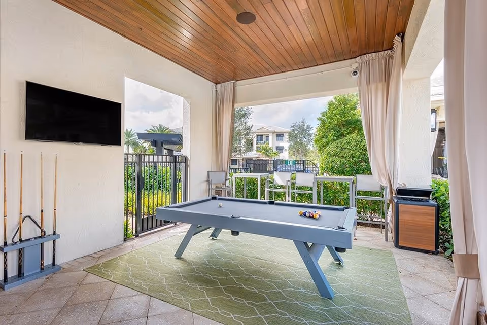 Covered outdoor game area with a pool table, mounted TV, seating and view of the landscaped courtyard.