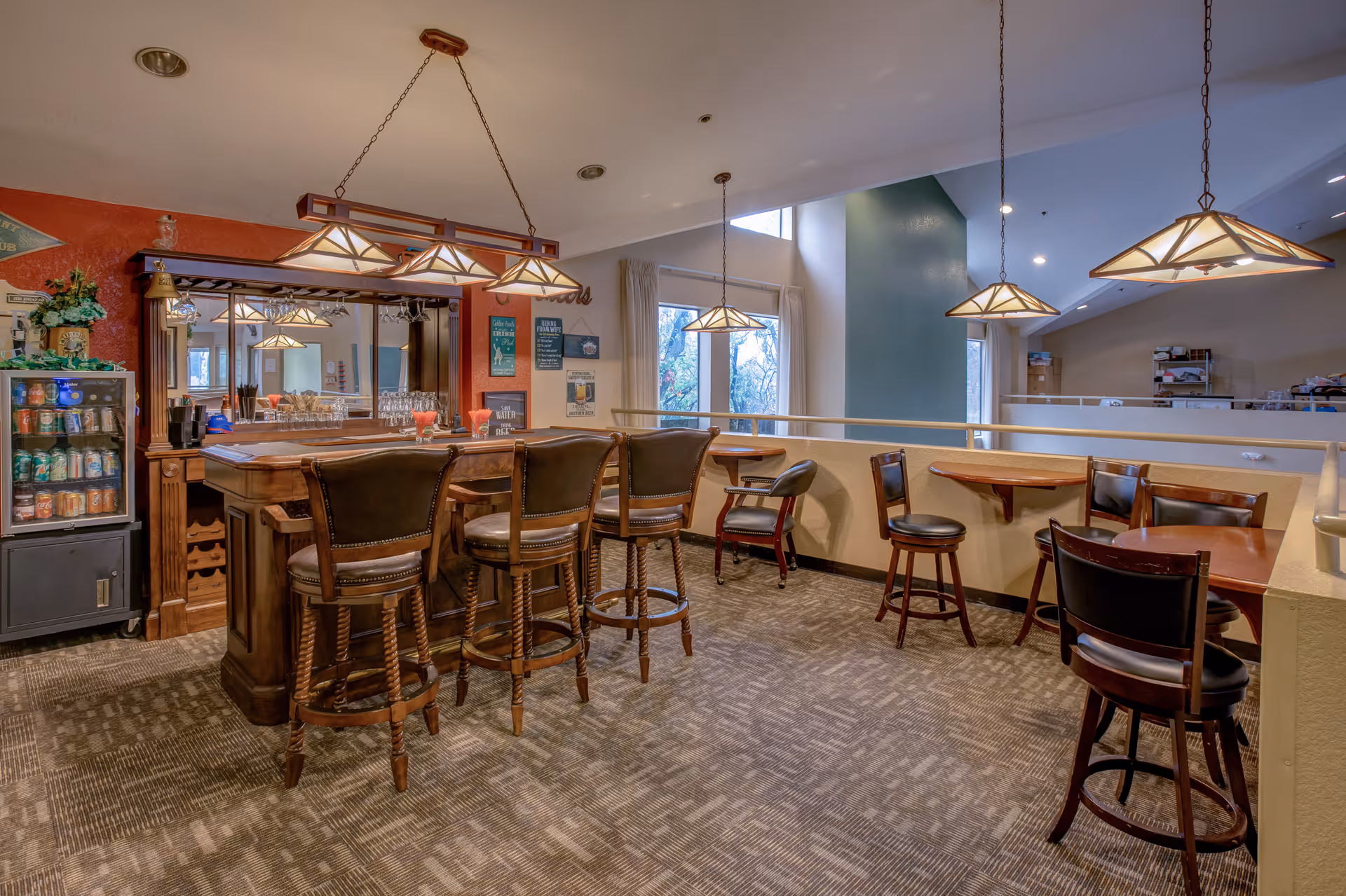 A communal bar and seating area with a wooden bar, barstools, pendant lights, and small tables in a retirement community.
