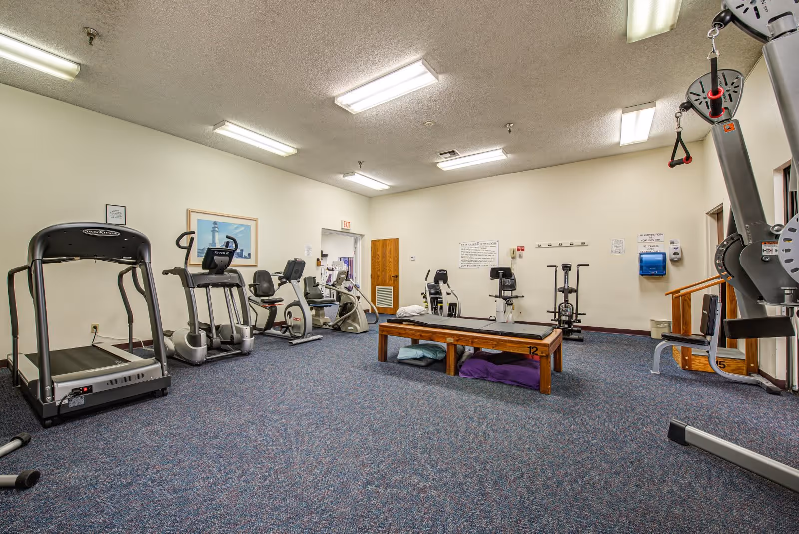 A fitness room in Westmont of Riverside featuring various exercise equipment including treadmills, stationary bikes, and weight machines. The room has blue carpet flooring, white walls, fluorescent ceiling lights, and a wooden bench in the center with pillows underneath. There is a framed picture on the wall and an exit door in the background.