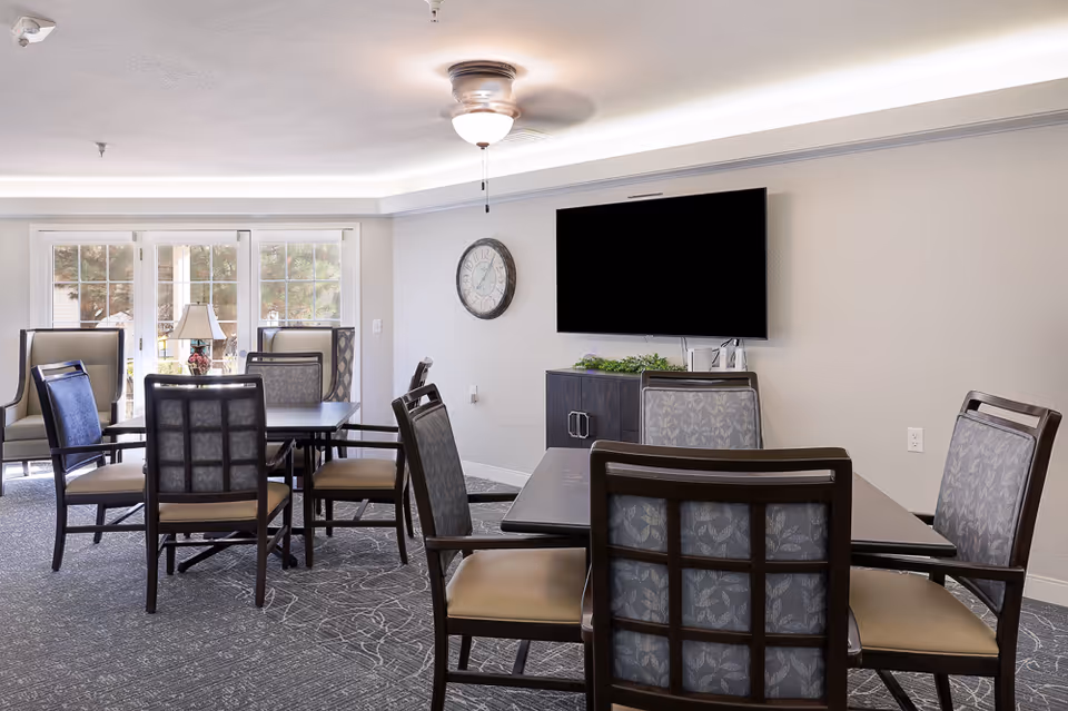 A well-lit common area in Homestead Assisted Living of Bethany featuring two tables surrounded by cushioned chairs with patterned backs. A large flat-screen TV is mounted on the wall above a cabinet, and a round wall clock is visible. French doors with glass panes lead outside, and a ceiling fan with a light fixture is mounted on the ceiling.