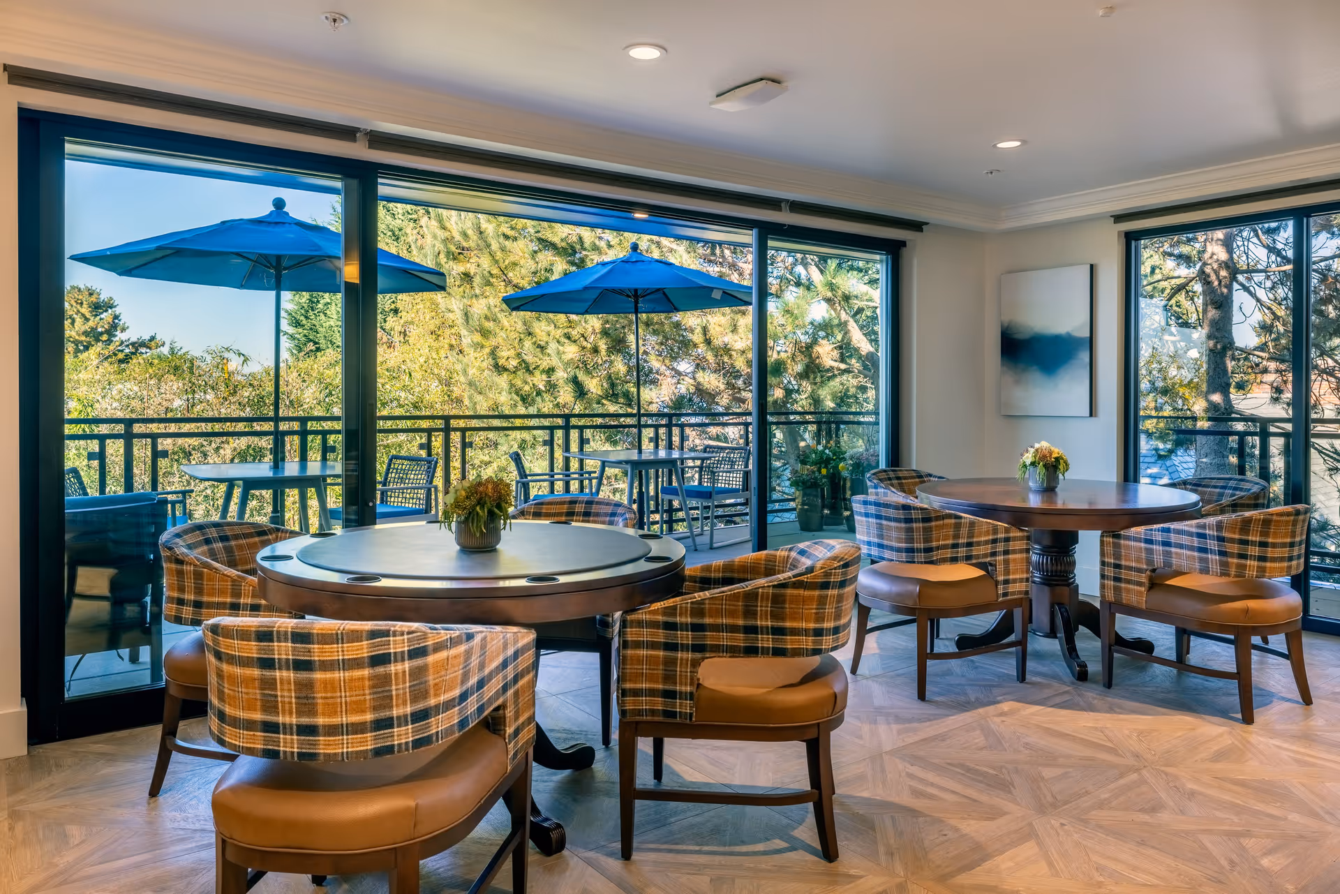 A bright dining area with two round wooden tables surrounded by plaid upholstered chairs. Each table has a small flower arrangement. Large glass sliding doors open to an outdoor patio with tables, chairs, and blue umbrellas. Trees and greenery are visible outside.