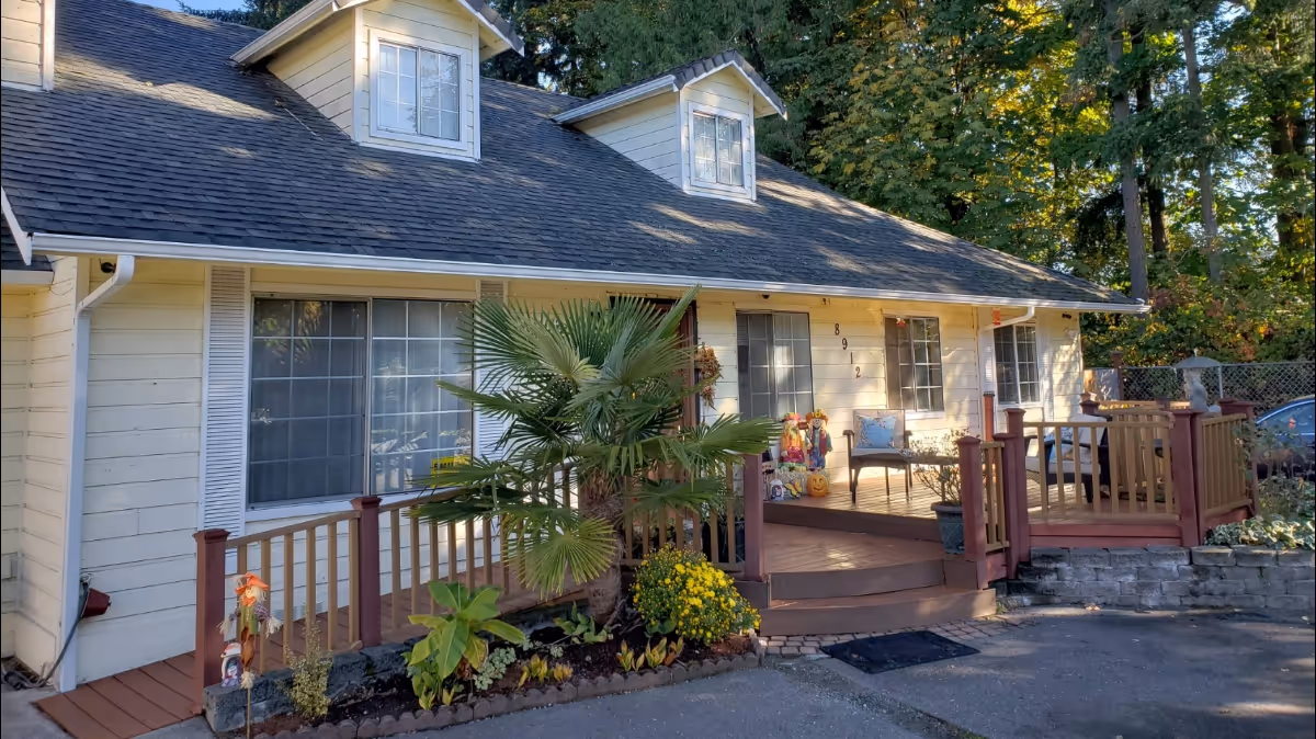 Front exterior view of a single-story house with a wooden porch, potted plants, and outdoor seating. The house has a sloped roof with two dormer windows and is surrounded by trees and greenery.